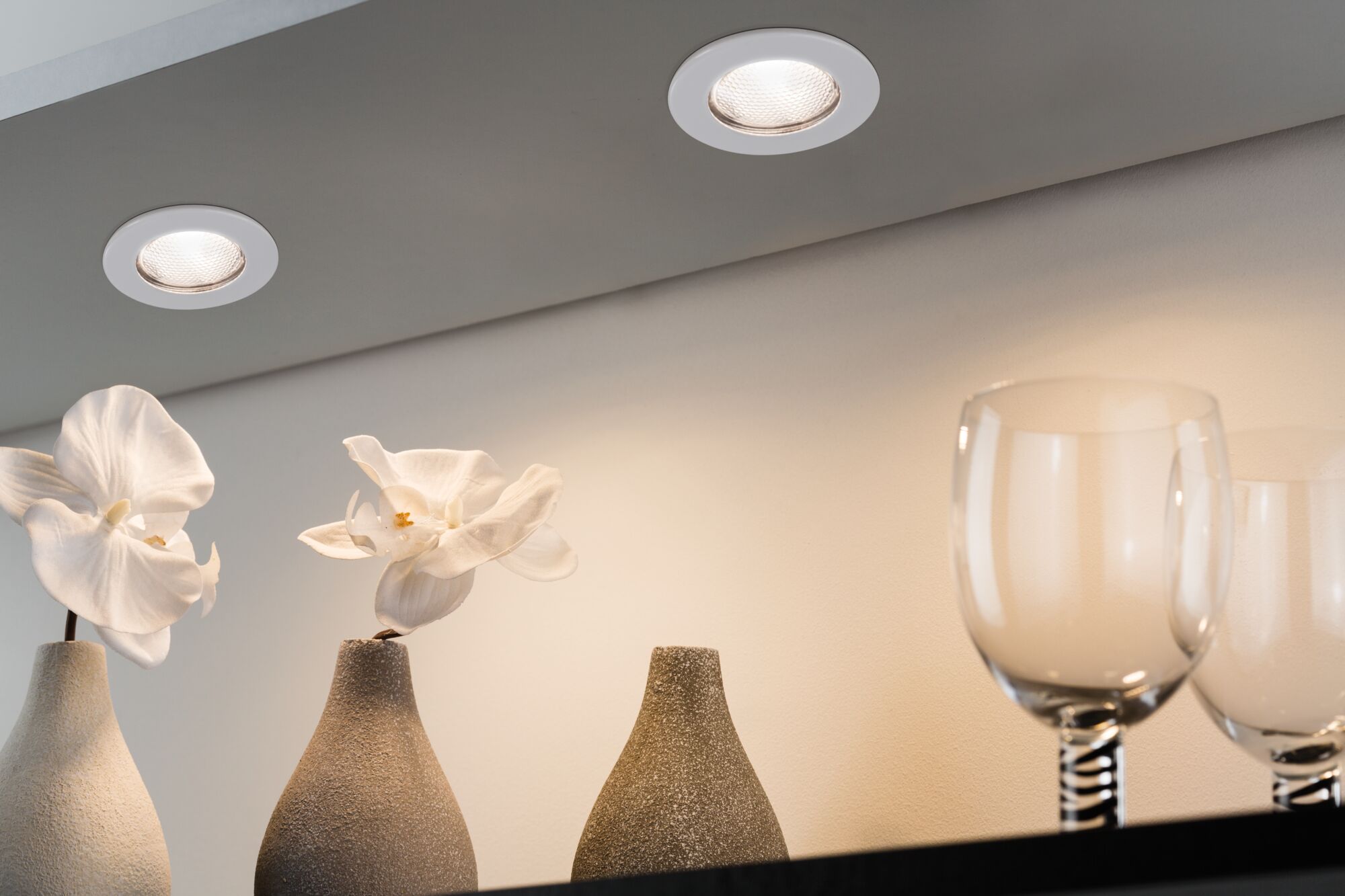 Deux spots LED encastrés blancs au plafond éclairent des orchidées blanches dans des vases en céramique grise et des verres.