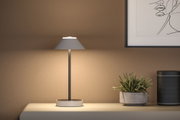 Moderne Tischlampe mit grauem Stoffschirm und Metallfuß auf beigem Sideboard neben Pflanze und Lautsprecher