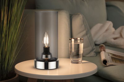 Modern table lamp with clear glass cylinder and chrome base on white side table next to glass of water