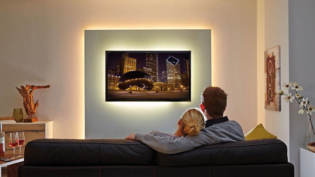 Paar auf schwarzem Ledersofa vor Wand mit indirekter LED-Beleuchtung und Fernseher im Wohnzimmer.