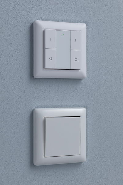 Twee witte kunststof wandschakelaars op blauwe muur, boven met LED-indicator, onder klassiek.