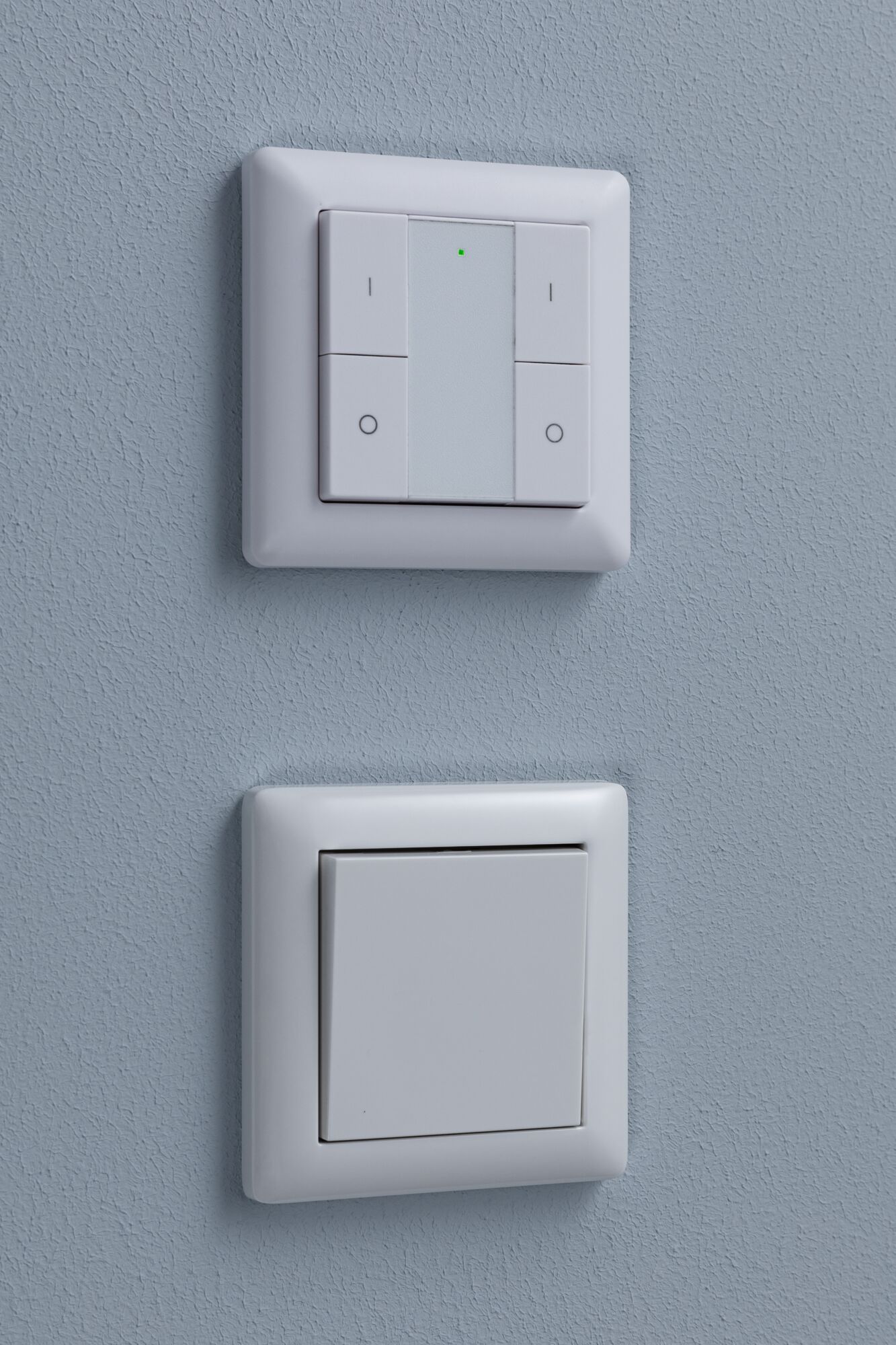 Twee witte kunststof wandschakelaars op blauwe muur, boven met LED-indicator, onder klassiek.