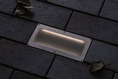 Rectangular stainless steel recessed light with mesh cover in dark pavement for outdoor lighting.