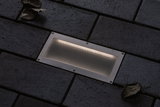 Rectangular stainless steel recessed light with mesh cover in dark pavement for outdoor lighting.