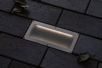Applique encastrée rectangulaire en acier inoxydable avec grille sur sol pavé sombre pour éclairage extérieur.