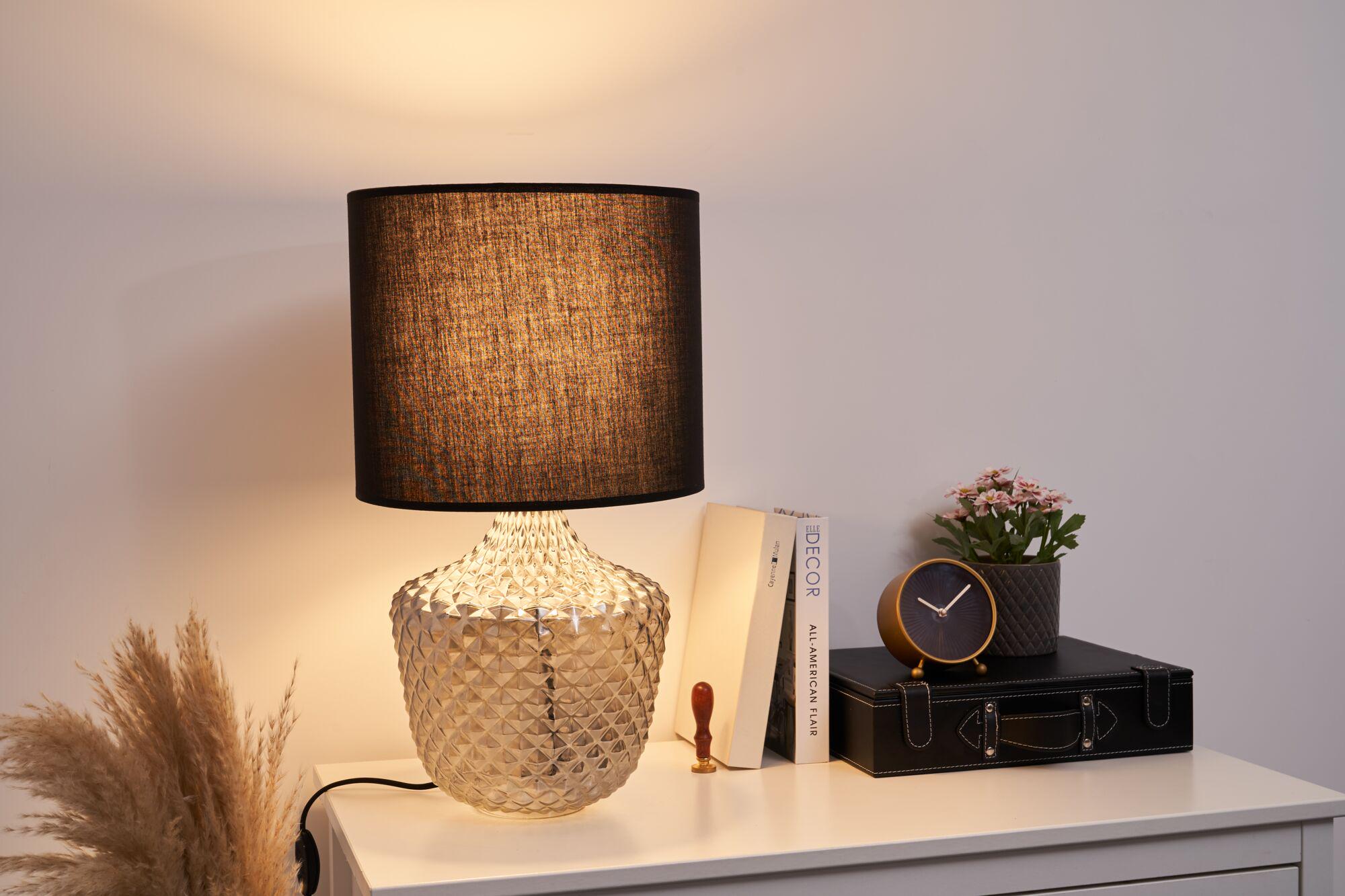Table lamp with textured glass base and black fabric shade on white bedside table with decor.
