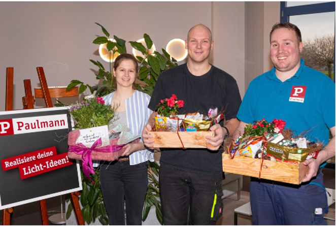 Drei Personen halten Geschenkkörbe mit Pflanzen und Snacks vor Paulmann Werbetafel für Lichtideen.