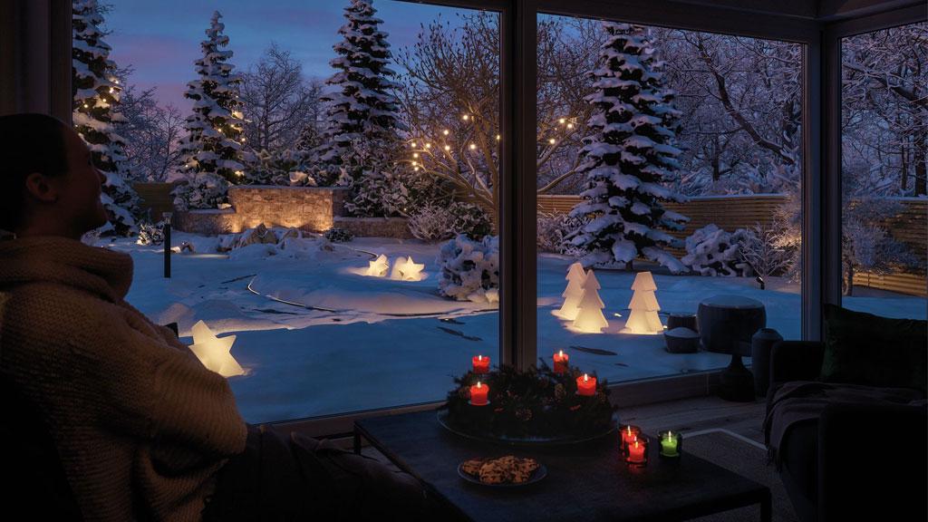 Wintergarten mit Adventskranz und Kerzen, draußen beleuchtete Weihnachtsdeko in Schnee mit Tannenbäumen.