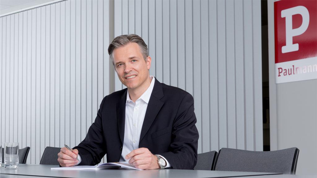 Mand i sort jakkesæt sidder ved bord med notesbog og pen i moderne kontor med Paulmann-logo