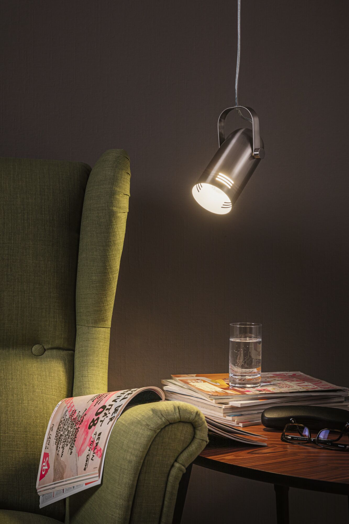 Moderner silberner LED-Hängeleuchte aus Metall über grünem Sessel und Holztisch mit Glas und Zeitschriften