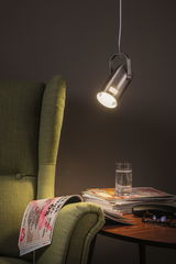 Moderner silberner LED-Hängeleuchte aus Metall über grünem Sessel und Holztisch mit Glas und Zeitschriften