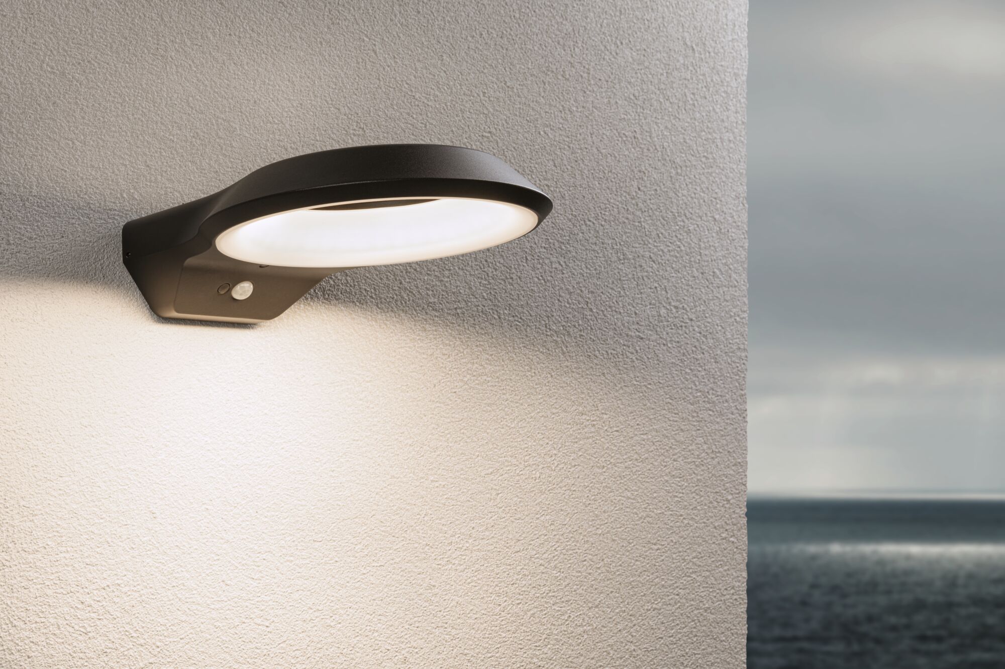 Schwarze Aluminium-Wandleuchte mit warmweißem LED-Ring und Bewegungsmelder an Außenwand mit Meerblick