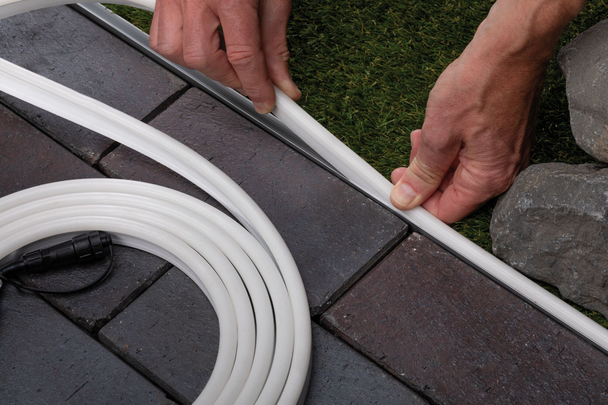Witte flexibele kabelmantel wordt langs rand tussen bestrating en gras gelegd voor buitenverlichting.