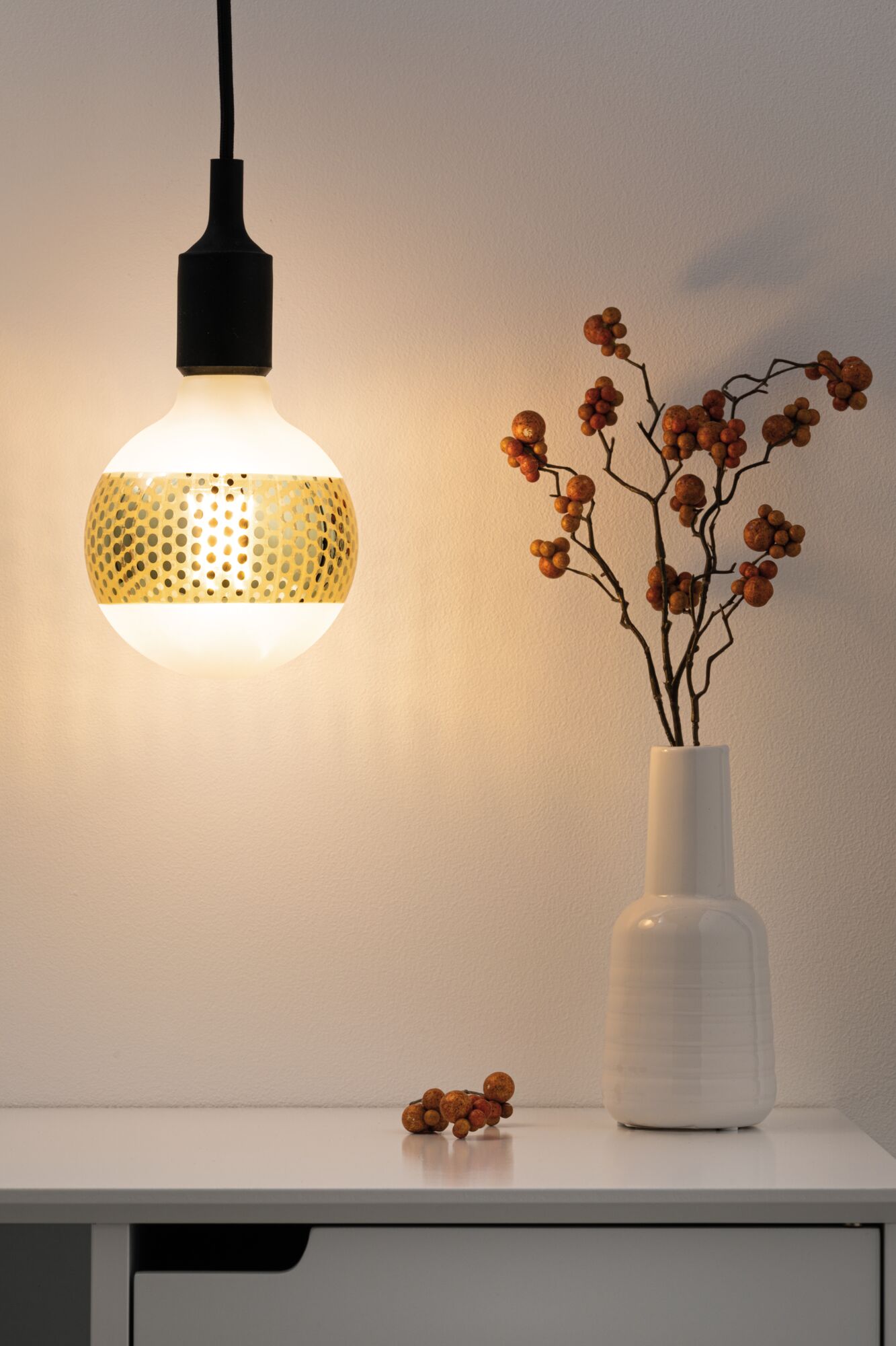 Hangende LED-lamp met goudkleurig geperforeerd patroon en zwarte kabel boven witte tafel met vaas en decoratie.