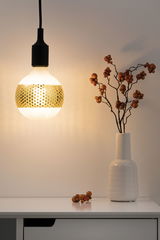 Hangende LED-lamp met goudkleurig geperforeerd patroon en zwarte kabel boven witte tafel met vaas en decoratie.