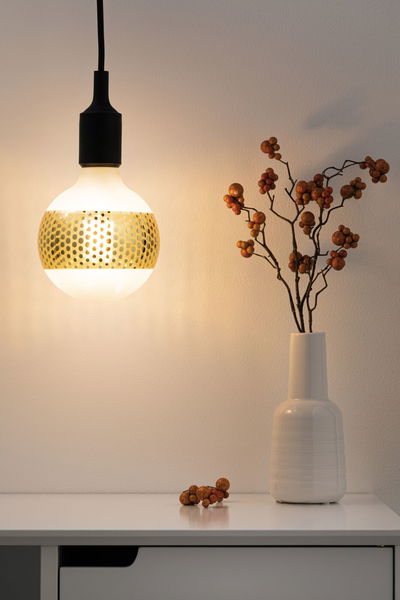 Hangende LED-lamp met goudkleurig geperforeerd patroon en zwarte kabel boven witte tafel met vaas en decoratie.