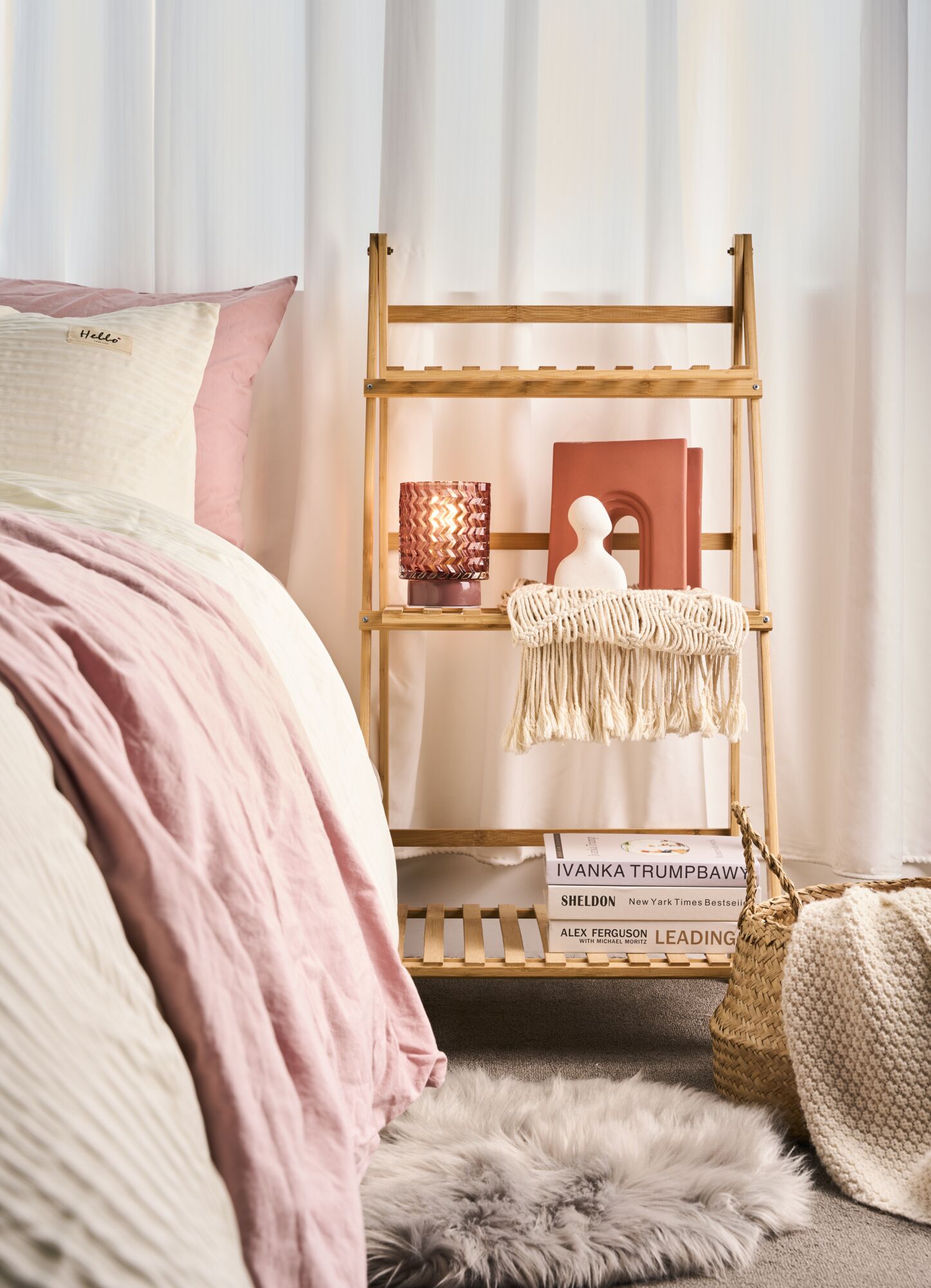 Bambusregal mit dekorativer Lampe, Büchern und Makramee in gemütlichem Schlafzimmer mit rosa Bettwäsche