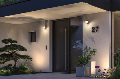 Moderne husfacade med sort dør, grå væglamper og dekorative planter ved indgangen.