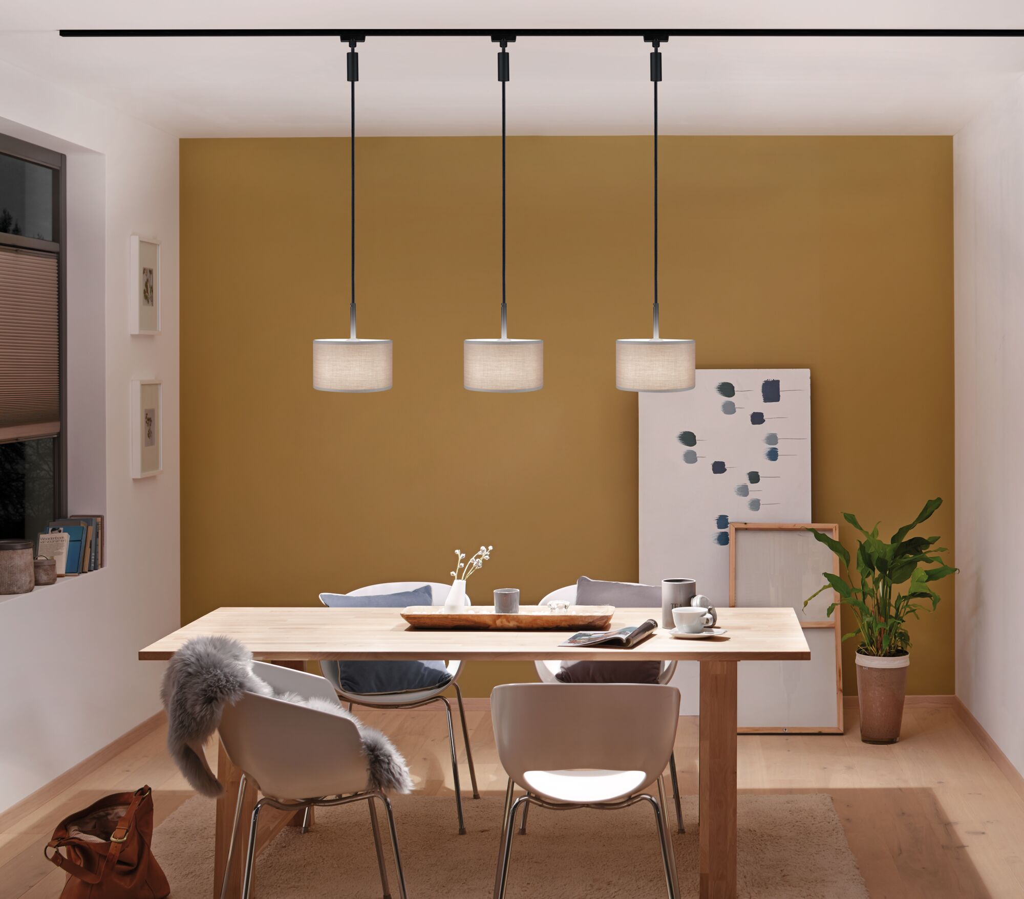 Drie moderne hanglampen met beige stoffen kap boven lichte houten eettafel in kamer met mosterdgele muur.