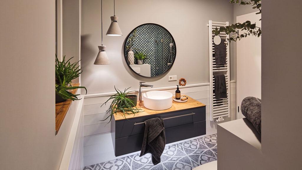 Modern bathroom with round mirror, wooden countertop, gray cabinet, and stylish lighting fixtures.