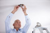 Older man installing round brushed stainless steel recessed spotlight on white ceiling, ladder nearby