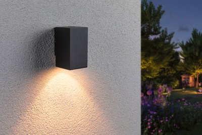 Black metal outdoor wall light with warm white illumination on textured wall in garden at dusk