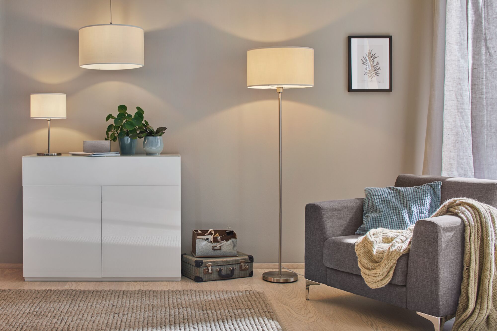 Moderne woonkamer met grijze stoffen fauteuil, wit dressoir en drie crèmekleurige lampen voor sfeerverlichting