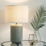 Lampe de table avec base en céramique grise et abat-jour en tissu blanc sur table en verre à côté d'un vase en verre avec feuille verte