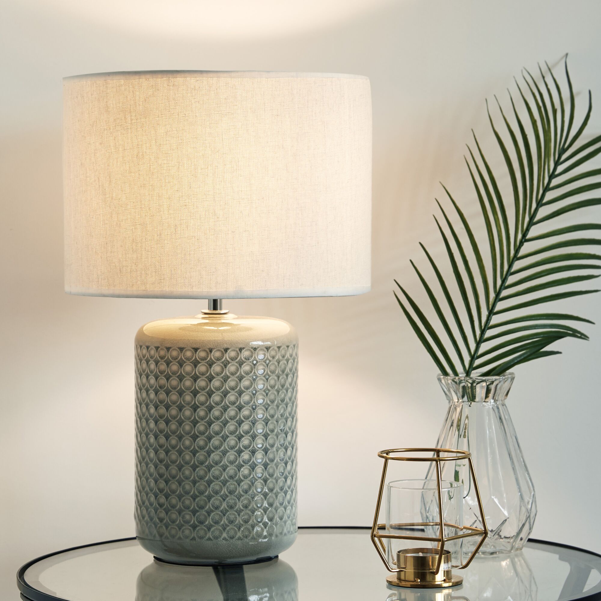 Lampe de table avec base en céramique grise et abat-jour en tissu blanc sur table en verre à côté d'un vase en verre avec feuille verte