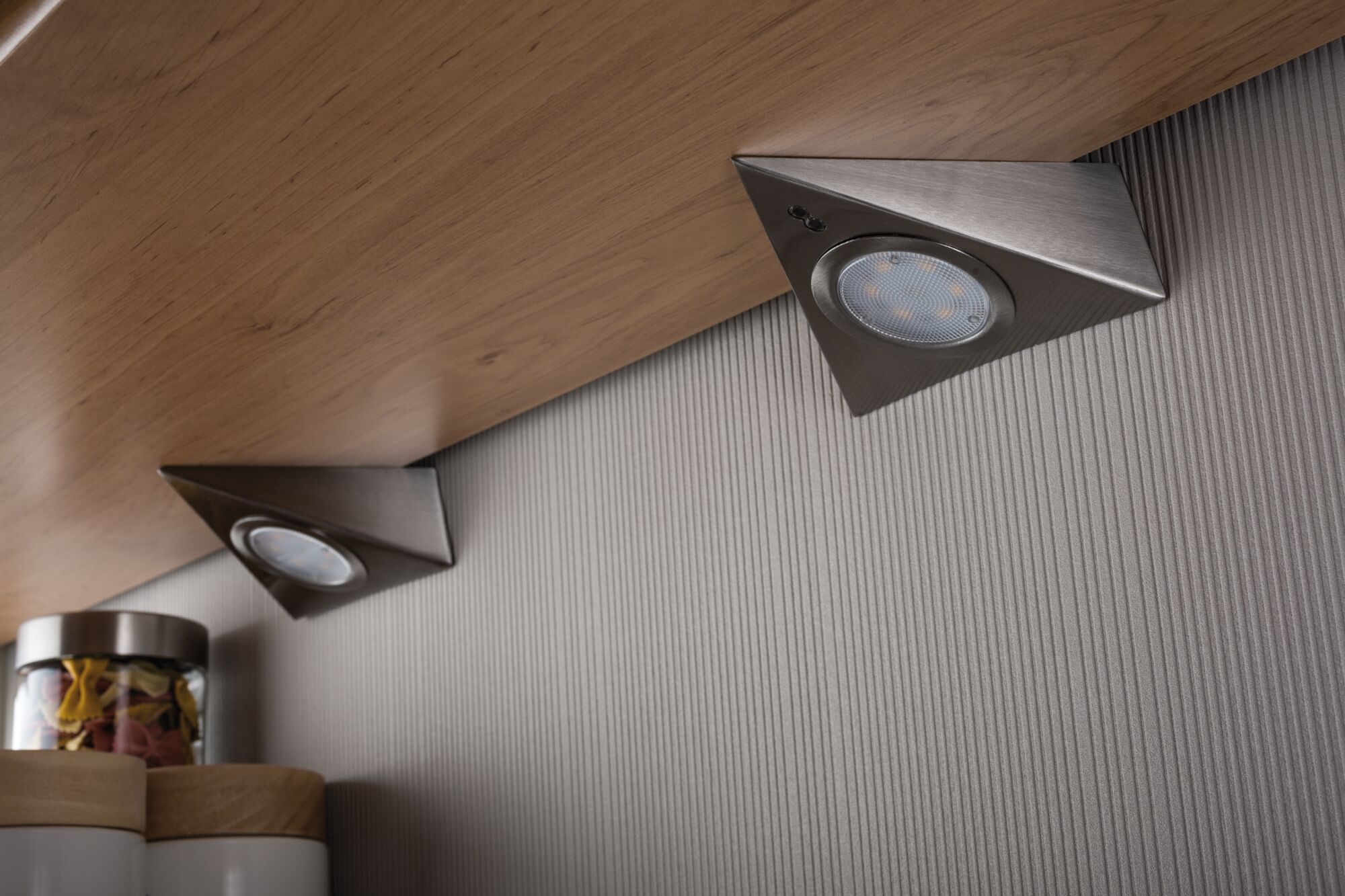 Dreieckige LED-Unterbauleuchten aus gebürstetem Edelstahl an Holzküche mit grauer Wand.