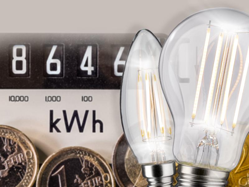 Zwei LED-Glühbirnen vor einem Stromzähler und Euro-Münzen symbolisieren Energieeinsparung und Kosten.