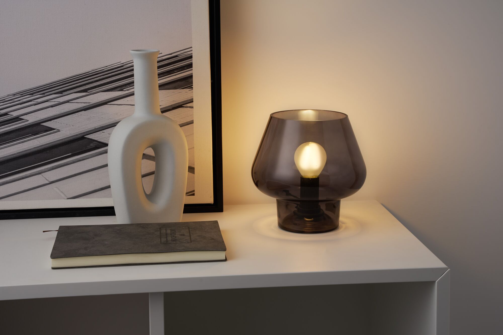 Modern table lamp with smoked glass and visible bulb on white shelf next to white decorative vase.
