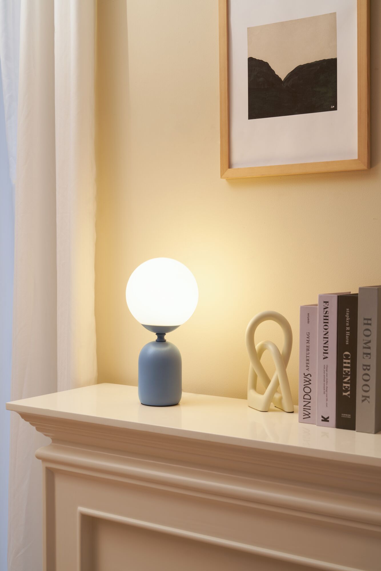 Moderne tafellamp met wit glas en grijze keramische voet op crèmekleurige schoorsteenmantel met boeken.