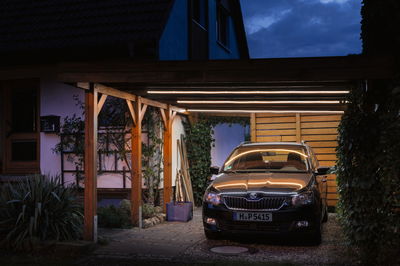 Wooden carport with warm white LED light strips illuminating black car at dusk in garden