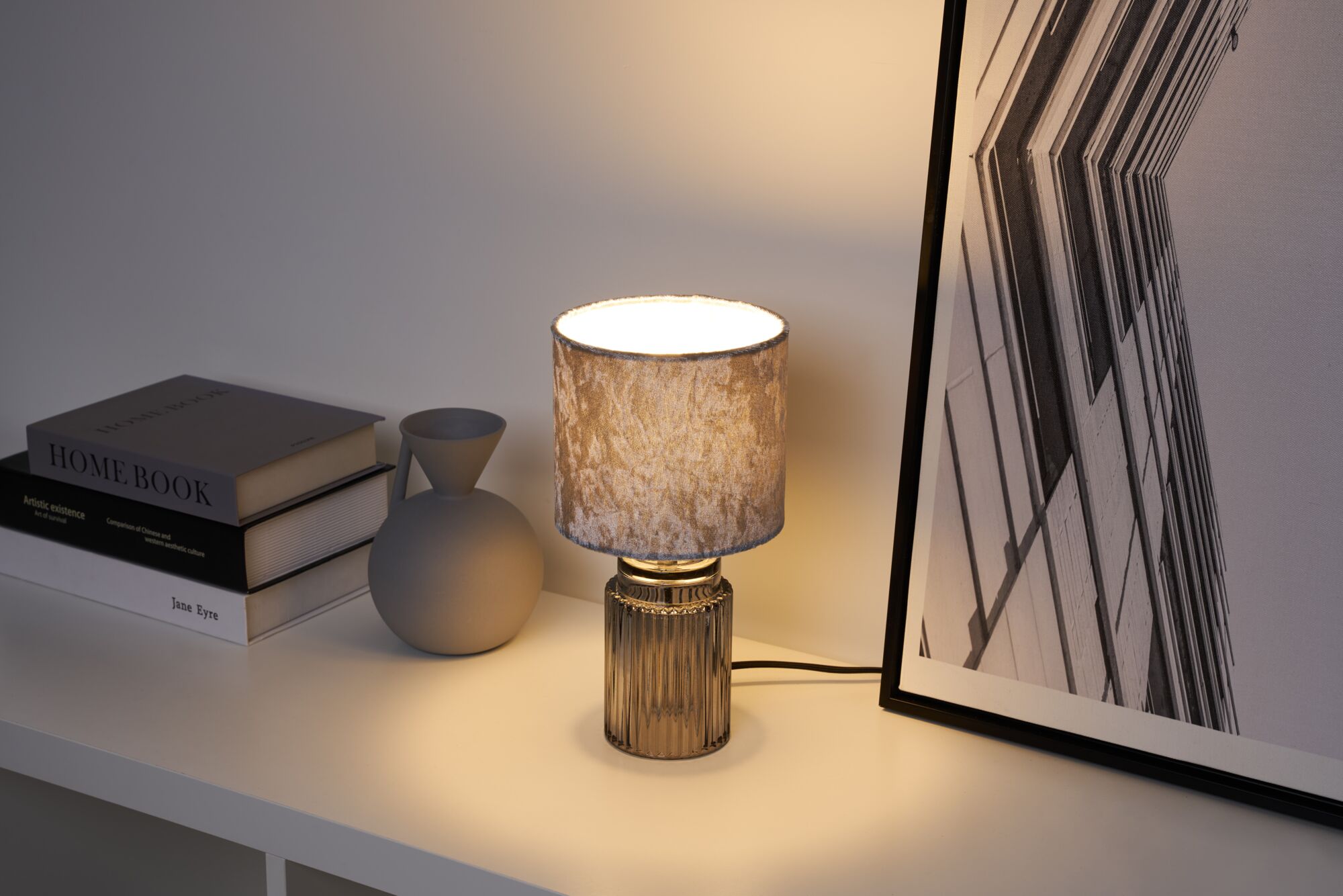 Moderne Tischlampe mit grauem Stoffschirm und geriffeltem Glasfuß auf weißem Regal mit Deko und Büchern