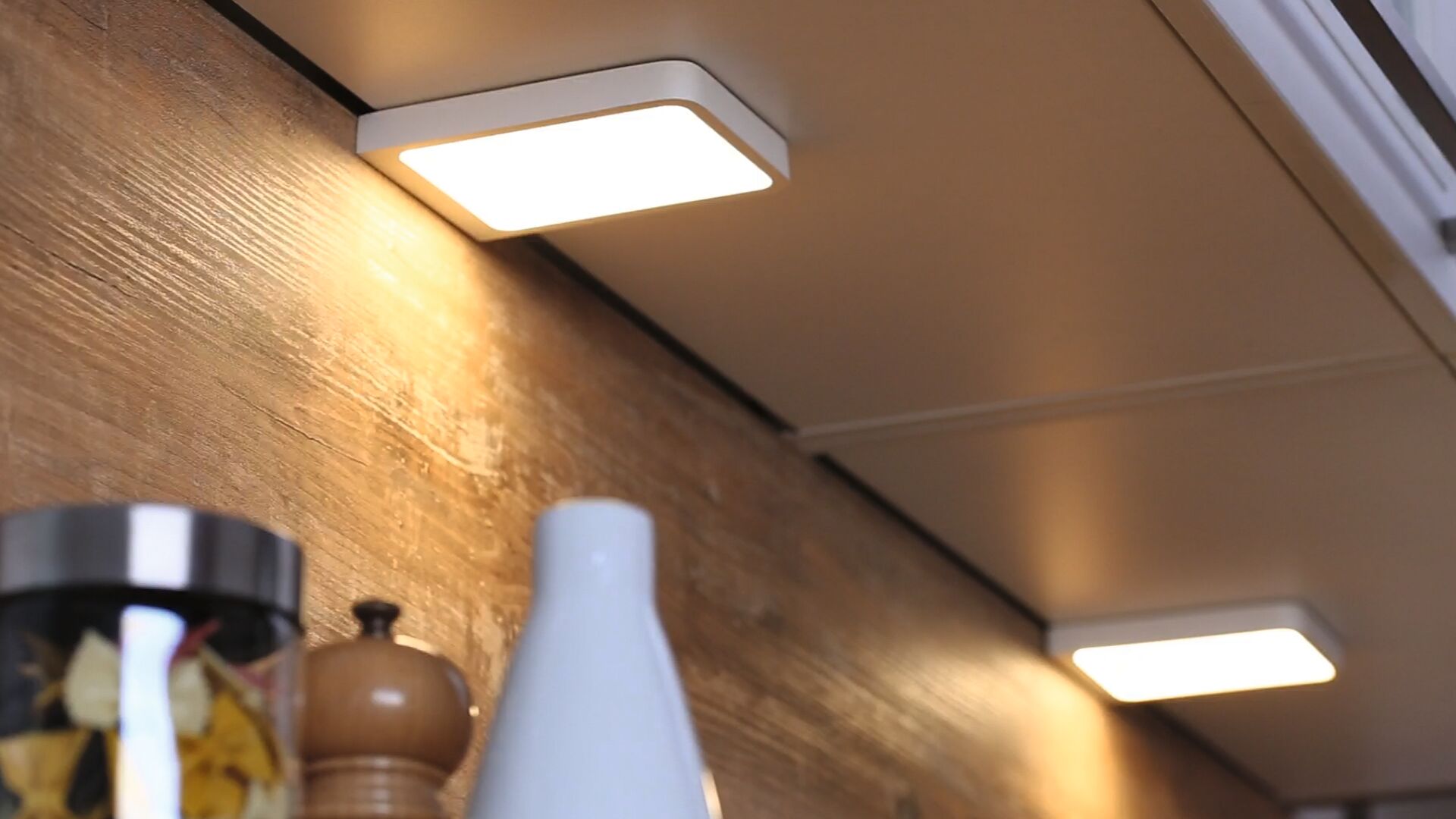 Square white LED under-cabinet light illuminating wooden kitchen backsplash with modern design