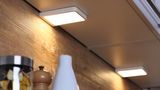 Square white LED under-cabinet light illuminating wooden kitchen backsplash with modern design