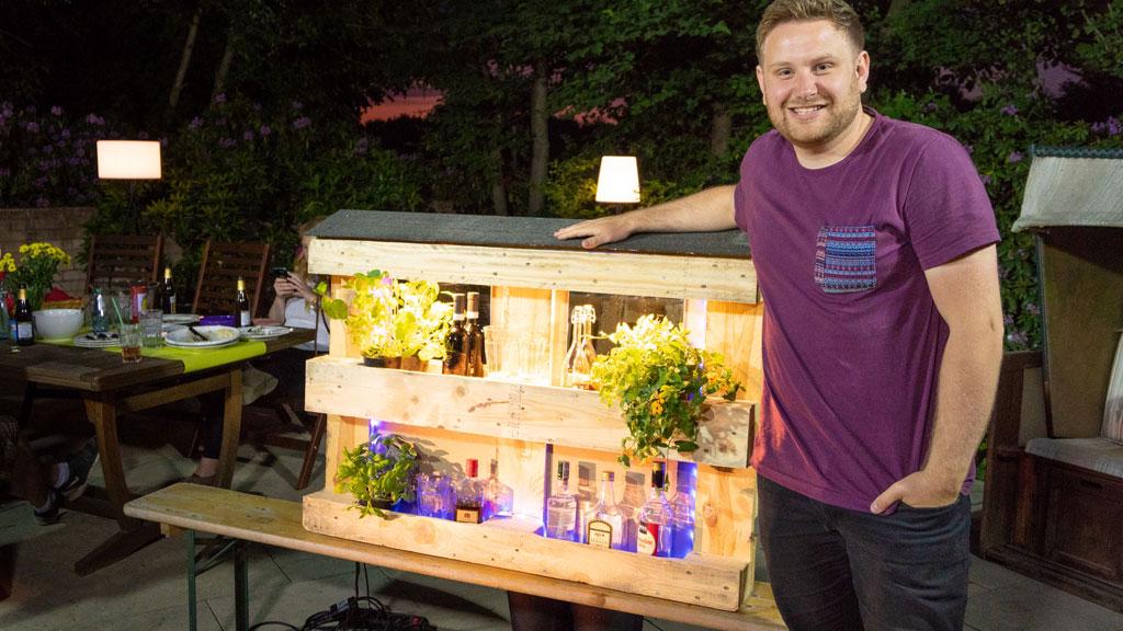Mann neben beleuchtetem Holzregal mit Pflanzen und Flaschen im Garten bei Abendlicht, Outdoor-Dekoration.