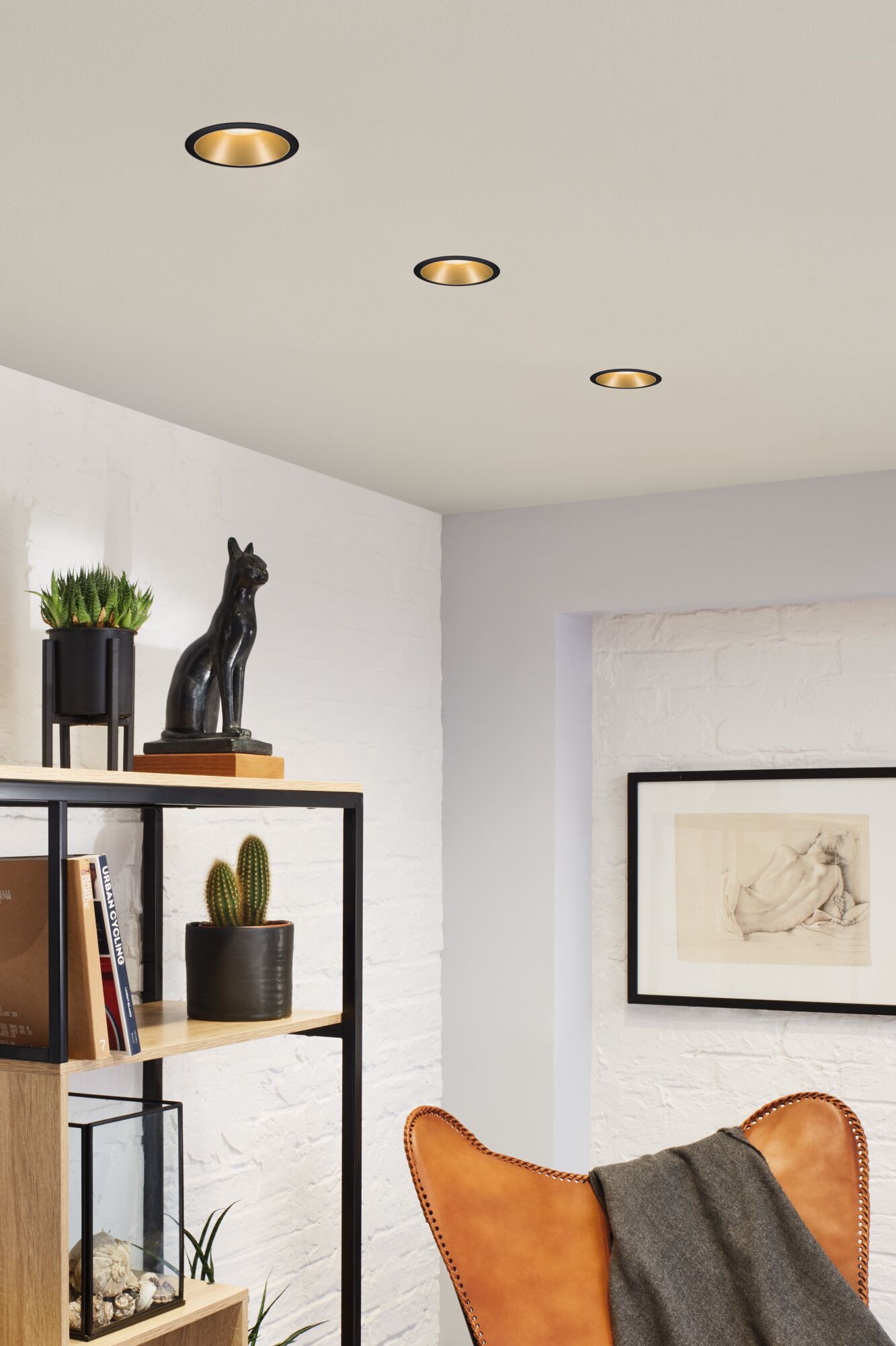 Three black recessed spotlights with gold interior illuminate modern living room with leather chair and plants.