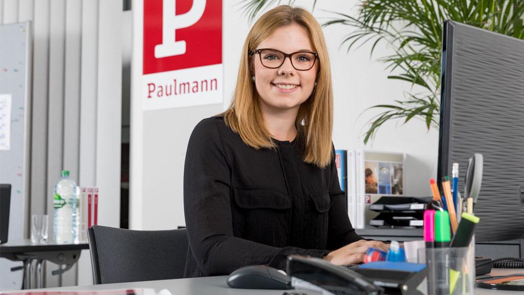 Junge Frau am Schreibtisch in modernem Büro mit Paulmann-Logo im Hintergrund, Arbeitsumgebung.