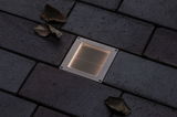 Recessed square LED floor light in stainless steel with warm white glow on dark pavement surface.
