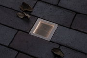 Recessed square LED floor light in stainless steel with warm white glow on dark pavement surface.