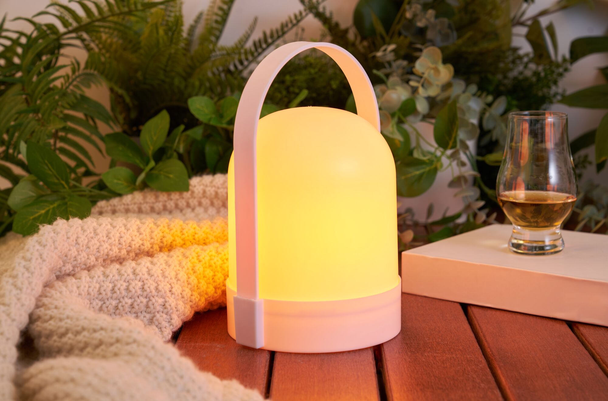 White portable LED light with warm glow on wooden table beside glass and knitted blanket
