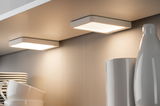 Two white square LED under-cabinet lights illuminating kitchen shelf with dishes and vases.