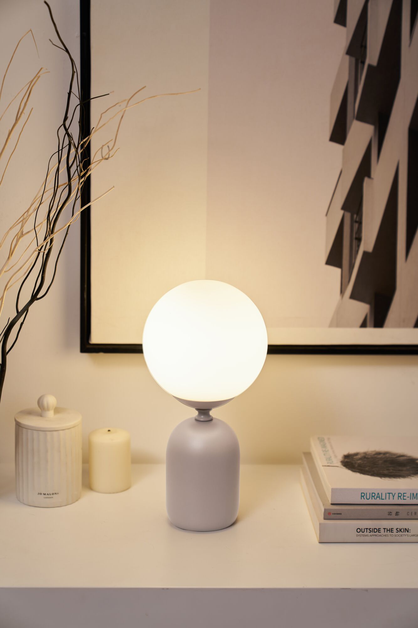 Lampe de table moderne avec verre blanc et base en céramique grise sur buffet blanc avec déco et livres.