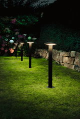 Four black LED garden lights with warm white glow on green lawn by stone wall and colorful flowers.
