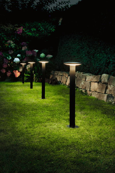 Vier schwarze LED-Gartenleuchten mit warmweißem Licht auf grünem Rasen neben Steinmauer und Blumen.