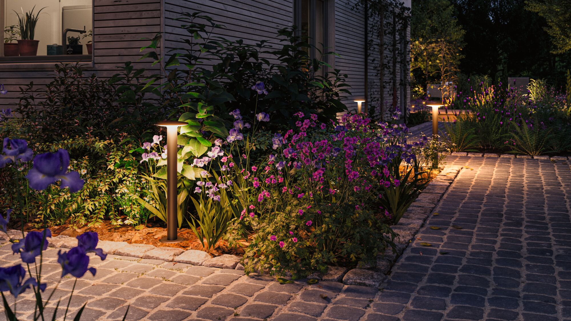 Moderne braune Außenleuchte aus Metall beleuchtet Gartenweg mit bunten Blumen bei Nacht