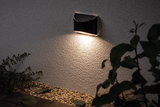 Black metal LED wall light with warm white illumination on exterior wall above gravel bed, garden lighting