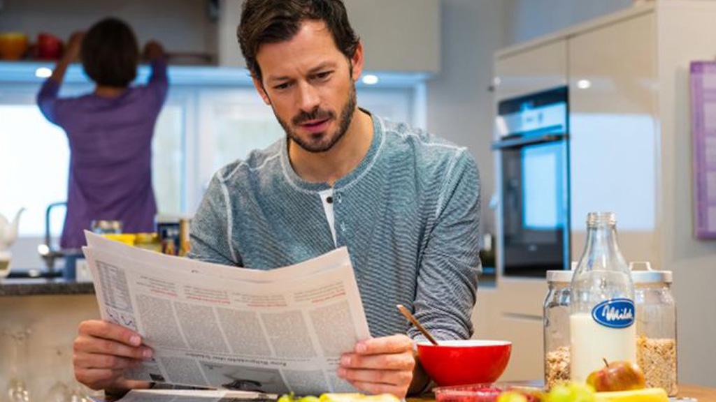 Mann liest Zeitung am Küchentisch mit Milchflasche, Müsligläsern und roter Schüssel im modernen Raum